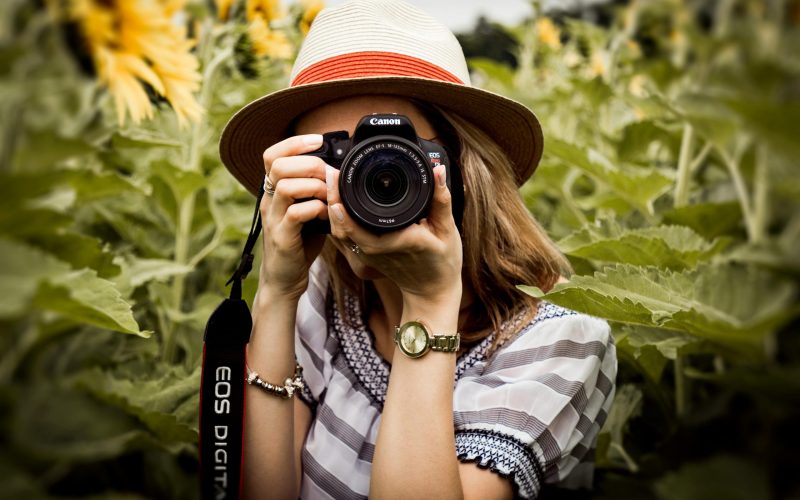 Woman with hat photographing sunflowers in a summer field using a Canon camera.