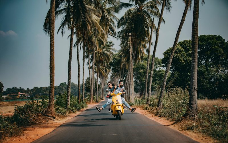 A joyful couple rides a yellow scooter through a scenic palm-lined road, capturing the essence of tropical travel and adventure.