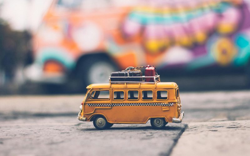 Close-up of a toy Volkswagen van with luggage, set against a colorful street backdrop, evoking summer travel vibes.